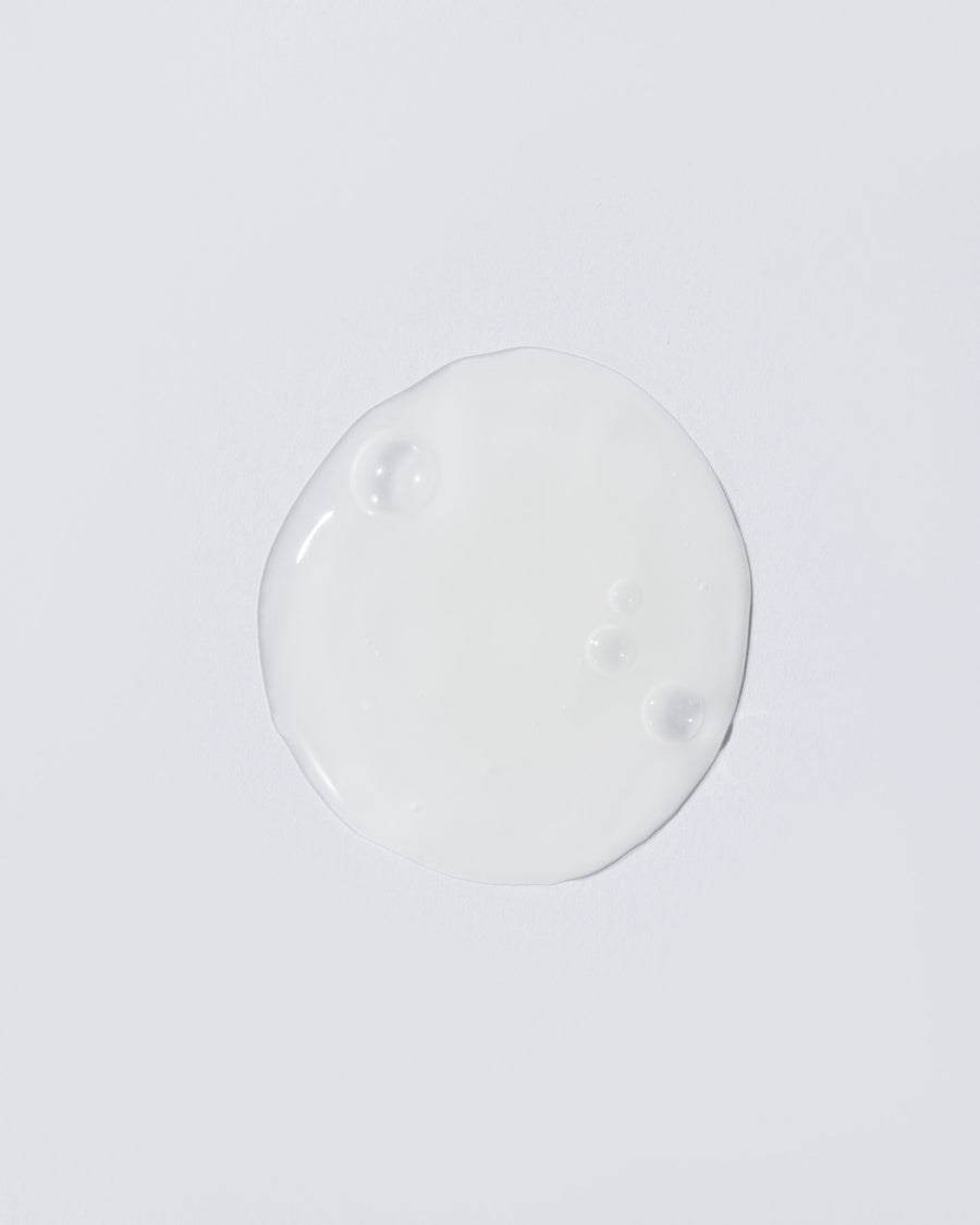 A glossy, translucent white circular droplet of cosmetic cream or gel centered on a solid light gray background.