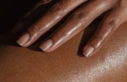 Crop anonymous African American female touching bare leg with hand while sitting on black background in room with bright sunlight