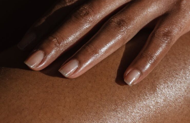 Crop anonymous African American female touching bare leg with hand while sitting on black background in room with bright sunlight
