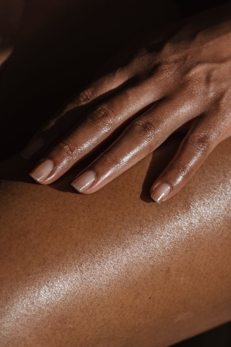 Crop anonymous African American female touching bare leg with hand while sitting on black background in room with bright sunlight
